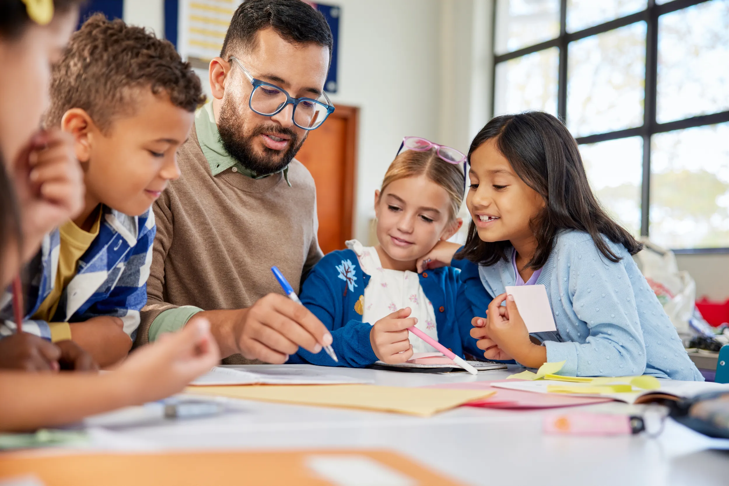 Male teacher with glasses helping a diverse group of young students with a classroom activity, all gathered around a table and smiling as they work together.
