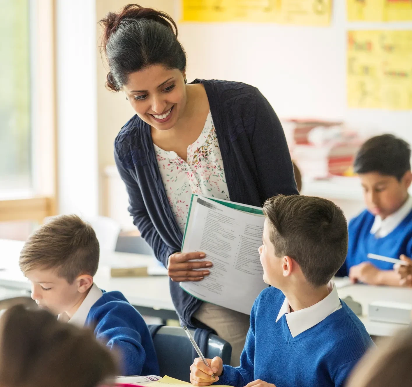 Smiling female teacher standing beside a student’s desk, holding papers and talking with a boy in a blue school uniform while other children work in the classroom.
